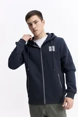 Campera con capucha de algodón y poliéster, color blanco, con cierre frontal negro y cordones negros en la capucha. Presenta un pequeño parche cuadrado negro con texto blanco en el pecho izquierdo y cierres negros en los bolsillos laterales.