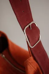 Bolso tipo hobo de gamuza color naranja quemado, con correa ancha de tela color bordó.