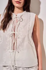 Blusa sin mangas de algodón color blanco off white, con cuello redondo y cierre frontal mediante cordones para atar. Presenta delicados detalles de bordados tonales e inserciones de encaje vertical en el frente.