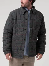 Chaqueta de hombre con diseño acolchado, confeccionada en lana Harris Tweed con patrón de tartán en tonos grises. Presenta cuello camisero, cierre frontal con botones de imitación cuero, bolsillos frontales y relleno térmico de fibras recicladas.