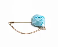 Pin para el ojal con diseño de rosa de satén en color celeste, montado sobre una aguja metálica dorada con cadena decorativa y detalle de hoja metálica.