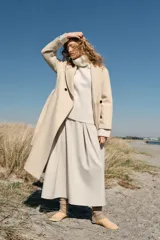 Abrigo largo de lana en color beige, con cuello de solapa, cierre cruzado con botones y cinturón ajustable del mismo tejido. Presenta mangas largas con detalle en los puños y bolsillos de vivo en el delantero.