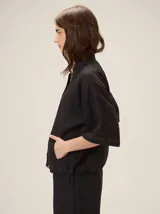 Chaqueta de lino y rayón color tostado, de corte holgado, mangas cortas amplias, cuello mao y cierre frontal oculto con cremallera. Presenta pespuntes a contratono en las costuras y cordón ajustable en el bajo.