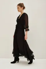 Vestido midi negro de estilo romántico, confeccionado en tejido semitransparente con detalles de encaje en el cuello, la cintura y el ruedo. Presenta mangas tres cuartos con terminación en encaje, escote en V con volados y un lazo de ajuste en la cintura.