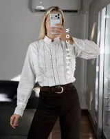 Camisa blanca de estilo romántico con cuello alto, cierre frontal de botones y detalles de encaje y alforzas verticales. Presenta mangas largas con puños decorados con encaje.