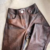 Pantalón de cuero vacuno en tono marrón con efecto desgastado, de corte recto y tiro medio. Cuenta con bolsillos delanteros tipo vaquero, trabillas para cinto y un botón frontal irregular de guampa.