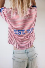 Camiseta oversize de rayas horizontales rojas y blancas, con cuello redondo y mangas cortas. Presenta un estampado de texto azul en el pecho y estrellas azules en las mangas.