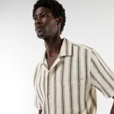 Camisa de manga corta color blanco con diseño de rayas verticales oscuras, cuello con muesca, cierre frontal con botones y bolsillo aplicado en el pecho.