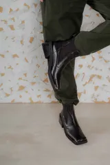 Botas tipo chelsea de cuero color marrón oscuro con acabado brillante. Presentan punta cuadrada con detalle de puntera metálica, elásticos laterales y taco cuadrado bajo.