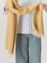 Bufanda de gran tamaño en color mostaza con diseño de tejido en espiga y flecos en los extremos. Incluye una etiqueta metálica decorativa en una de las esquinas.