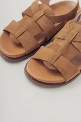 Sandalias planas de gamuza color marrón, con diseño de tiras cruzadas en el empeine y cierre con hebilla metálica dorada en el tobillo. Cuenta con plantilla de cuero y suela de goma con textura.