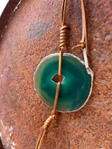 Collar con dije de piedra ágata blanca y marrón, con forma circular irregular y agujero en el centro. El dije está sujeto con un cordón fino de cuero marrón.