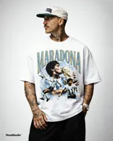 Remera blanca de algodón premium de alto gramaje, con estampa frontal que incluye imágenes de Diego Maradona besando la Copa del Mundo y jugando al fútbol, con el texto "MARADONA" en tipografía grande.