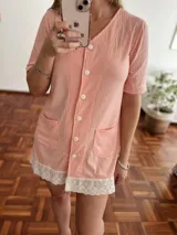 Camisola o bata corta de tejido acanalado color salmón, con cuello en V, manga corta y cierre frontal con botones blancos. El ruedo está decorado con un borde de encaje blanco.
