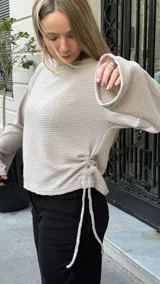 Buzo de punto color beige con textura acanalada, cuello redondo y mangas largas. Presenta un diseño ajustable en los laterales mediante tiras que permiten modificar el largo de la prenda.