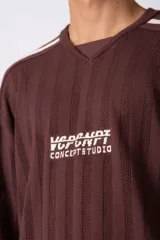 Remera de tejido de punto con textura calada, color crema con rayas verticales sutiles. Presenta cuello en V, mangas cortas con franjas laterales en color marrón oscuro y logo bordado en el centro del pecho.