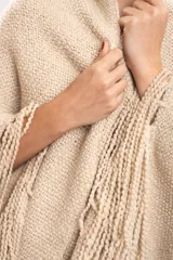 Poncho triangular tejido artesanalmente en telar manual, de lana merino color beige, con cuello alto y flecos largos en los bordes.