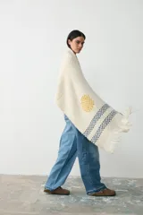 Chal tejido de punto en color crudo, con diseño artesanal que incluye un círculo amarillo y franjas horizontales en azul, con terminación de flecos en los extremos.