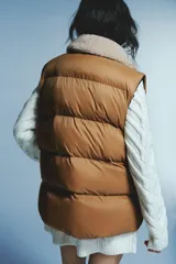 Chaleco acolchado de color tostado con diseño guateado, cuello de sherpa en tono crema, cierre frontal con cremallera y bolsillos frontales tipo parche.
