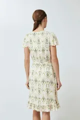 Vestido corto cruzado con estampado abstracto en tonos verde oliva, amarillo y rosa. Tiene escote en V, mangas cortas con volados y falda con volados en el ruedo. Se ata a la cintura con un lazo.