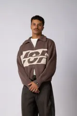 Campera tejida de punto con cierre frontal completo y cuello polo. Presenta un diseño de franja horizontal ancha en color crema con estampado gráfico oscuro sobre una base de color marrón. Posee puños y ruedo acanalados, con un pequeño parche de marca en el puño izquierdo.