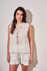 Blusa sin mangas de algodón color blanco off white, con cuello redondo y cierre frontal mediante cordones para atar. Presenta delicados detalles de bordados tonales e inserciones de encaje vertical en el frente.