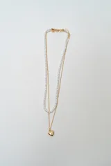 Collar doble de cadena dorada fina, una de ellas con perlas pequeñas y la otra con un dije de corazón dorado colgante.
