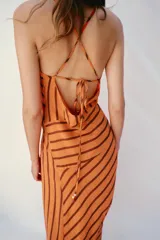 Vestido midi de color naranja con rayas diagonales marrones, escote fluido y tirantes finos cruzados en la espalda con abalorios. Presenta corte evasé con abertura frontal y forro interior.