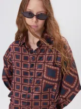 Camisa de seda con estampado geométrico de cuadrados en tonos rojizos y marrones. Presenta cuello camisero, cierre frontal con botones, bolsillo en el pecho y mangas largas con puños abotonados.