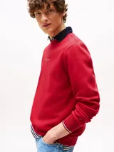 Buzo rojo de cuello redondo con ribetes distintivos, logo texturizado de Tommy Hilfiger en el pecho y logo bordado en la manga. El cuello es azul marino y se asoma un cuello de camisa a rayas. Los puños tienen ribetes distintivos.