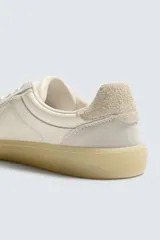 Zapatilla deportiva tipo barefoot confeccionada en piel color blanco roto con apliques de gamuza en tonos grises. Posee puntera redondeada y ancha, cierre mediante cordones al tono y suela de goma flexible en color caramelo con tecnología drop 0.