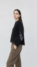 Sweater de lana color beige con calce holgado y cuello redondo. Presenta un bordado negro con diseño de arabescos en una de las mangas y detalles de rayas y puntos en contraste en el ruedo y los puños.