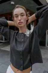 Chaqueta gris de punto con cuello redondo, manga larga y cierre frontal con botones. Detalle de flor blanca y negra en el frente. Tejido en rib.