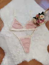 Conjunto de lencería compuesto por un bralette triangular y una tanga, confeccionados en algodón color rosado con detalles de encaje en tono crema. El bralette cuenta con breteles ajustables y el conjunto presenta terminaciones delicadas.