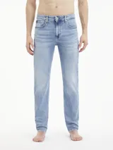 Jeans de mezclilla azul claro, tiro medio, con corte slim que se estrecha hacia el tobillo. Presentan un diseño de cinco bolsillos, bragueta con cremallera y herrajes de Calvin Klein, incluyendo una insignia de cuero en la parte trasera.