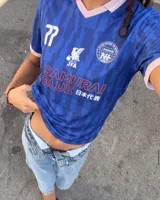 Camiseta de fútbol de manga corta en color azul con diseño estampado abstracto y cuello en V rosado con detalles blancos a juego con los puños. Presenta inscripciones y escudos deportivos en el pecho.