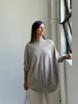 Poncho de tejido de punto color visón, con cuello alto y diseño de trenzas verticales. Presenta mangas cortas ajustadas y un corte holgado y redondeado en el ruedo.