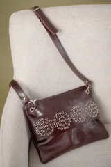 Sobre de cuero vacuno color bordó, con diseño frontal decorado con múltiples remaches metálicos circulares. Cuenta con correa larga ajustable y extraíble, cierre superior y bolsillo trasero con cierre.