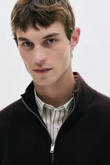 Buzo de punto color marrón oscuro (casi negro), de manga larga, con cuello alto y medio cierre metálico frontal. Presenta un tejido acanalado en el cuello y un tejido liso en el cuerpo.