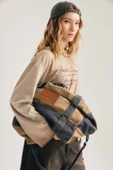 Bolso estilo duffel o bandolera de diseño bicolor, combinando lona en tono tostado y denim gris gastado. Cuenta con asas superiores, correa ajustable para el hombro y un parche de cuero marrón con el logotipo de la marca grabado en la parte frontal.