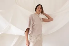 Top beige asimétrico de un solo hombro con mangas murciélago amplias, adornado con detalles de flecos delicados en el escote y en el ruedo inferior.