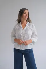 Blusa blanca de manga larga con diseño de rayas verticales y bordados florales en tono grisáceo. Presenta cuello camisero, cierre frontal con botones y puños elásticos.