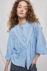 Blusa celeste de manga 3/4 con cuello mao, botonadura frontal y detalles calados en V con frunces. Presenta mangas con volumen y silueta fluida.