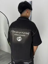 Camisa de manga corta color negro con efecto lavado, confeccionada en denim. Presenta costuras a la vista en color blanco y un estampado en la espalda con la frase 'Never give up on yourself, commit to the world' junto al logo de la marca Mozaiko.