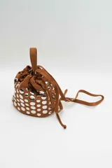 Cartera tipo bucket de gamuza color marrón, con diseño de tachas metálicas circulares en toda la superficie. Cuenta con cierre de cordón ajustable, asa superior y correa larga regulable.