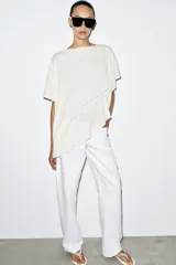 Top tipo capa blanco de punto fluido, con mezcla de viscosa, cuello bote, mangas cortas y bajo asimétrico.
