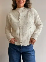 Cárdigan de punto color crema con motas de colores, diseño de trenzas en los laterales, cuello redondo y cierre frontal con botones. Posee puños y ruedo acanalados.