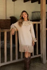 Vestido de punto color beige con diseño holgado, cuello redondo con volados de tela, mangas largas con puños con volados y ruedo decorado con flecos largos.