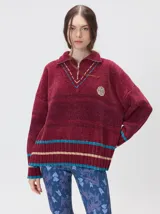 Pullover de textura aterciopelada en color bordó, con cuello polo y cierre frontal parcial. Presenta detalles de pespuntes decorativos en el cuello, franjas horizontales en tonos tierra y azul en el ruedo y puños, y un aplique bordado en el pecho. Posee un corte oversize y terminaciones acanaladas.
