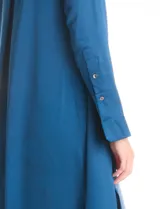 Túnica de satén color azul petróleo, con cuello a la base, cierre frontal con botones y mangas largas con puños acampanados. Presenta un diseño asimétrico con el largo de la espalda mayor al delantero.